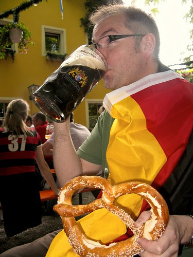 Geoff at a Munich beer garden drinking a litre glass of German beer with a German flag and a large Bretzn